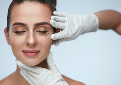 Eine Frau mit geschlossenen Augen. Sie hat markierte Linien unter einem Auge und wird von Händen in weißen medizinischen Handschuhen sanft am Kinn und an den Schläfen gehalten, was auf eine kosmetische Behandlung hinweist.