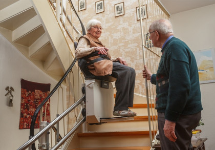 Ein älteres Paar in einem Haus. Eine Frau sitzt auf einem Treppenlift und fährt die Treppe hinauf, während ein Mann neben dem Treppenlift unten an der Treppe steht. Die Frau lächelt und schaut den Mann an.