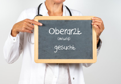 Eine Person in einem weißen Arztkittel hält ein kleines Tafelschild. Auf der Tafel steht in weißer Kreideschrift: "Oberarzt (m/w/d) gesucht". Die Person zeigt mit einem Finger auf das Schild.