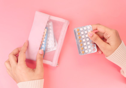 Eine Person hält eine Blisterpackung mit Tabletten in der einen Hand und greift mit der anderen Hand nach einer rosa Tasche, die ebenfalls Tablettenpackungen enthält. Der Hintergrund ist rosa.