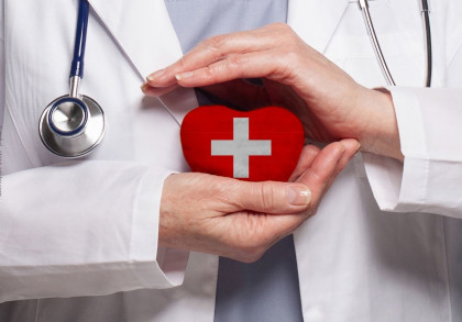 Nahaufnahme eines Arztes in weißem Kittel mit Stethoskop um den Hals, der ein rotes Herz mit einem weißen Kreuz darauf in den Händen hält. Die Szene symbolisiert medizinische Fürsorge und Herzgesundheit.