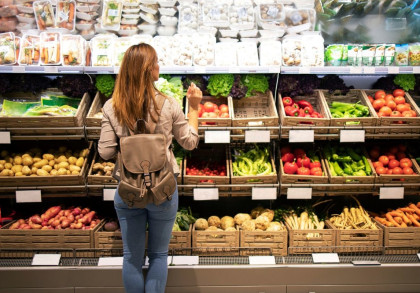 Eine Frau mit braunem Rucksack steht vor einem Gemüseregal in einem Supermarkt. Das Regal ist mit verschiedenen frischen Gemüsesorten wie Kartoffeln, Tomaten, Paprika und Karotten gefüllt.