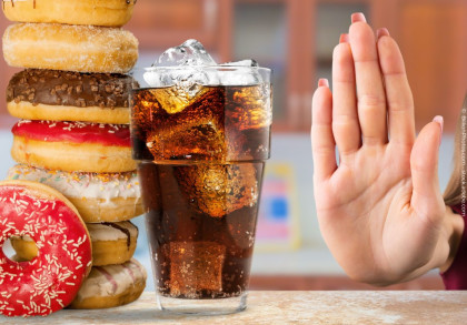 Eine Nahaufnahme von einem Stapel Donuts mit verschiedenen Glasuren und einem Glas Cola mit Eiswürfeln. Neben dem Glas ist eine Hand zu sehen, die eine "Stop"-Geste macht.