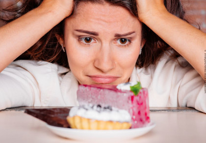 Frau stützt ihren Kopf auf ihre Hände und schaut besorgt auf ein Stück Kuchen und eine Torte auf einem Teller vor ihr.