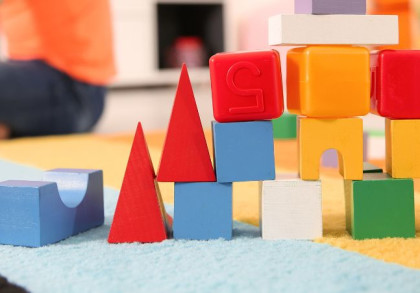 Bauklötze bilden einen Turm in einem Spielzimmer. Im Hintergrund sitzt ein Kind im Schneidersitz und schaut nach links.