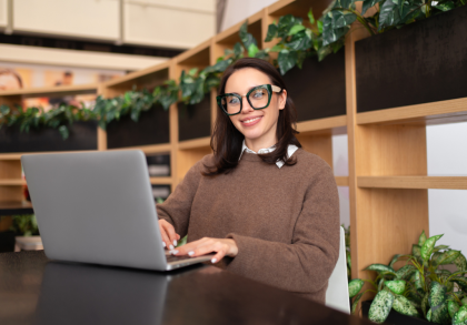 Frau mit großer grüner Brille sitzt lächelnd an einem Laptop in einem modernen, hellen Innenraum mit Holzregalen und Pflanzen im Hintergrund.