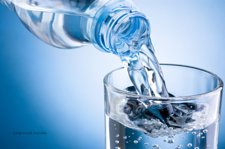 Eine Flasche Wasser wird in ein Glas gegossen vor einem blau HIntergrund.