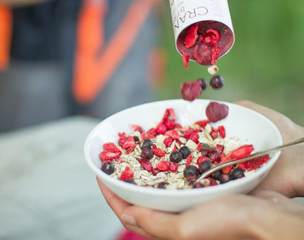 Eine Schüssel Müsli mit Haferflocken und roten Beeren wird gehalten. Aus einer Packung werden getrocknete Himbeeren und Blaubeeren in die Schüssel hinzugefügt. Eine Person hält die Schale mit einer Hand und hat einen Löffel in der anderen Hand.