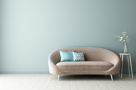Ein modernes Wohnzimmer mit einer taupefarbenen Couch, die sich vor einer mintgrün gestrichenen Wand befindet. Zwei Dekokissen in Türkis und mit geometrischem Muster zieren die Couch.