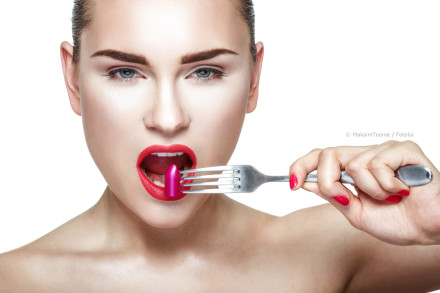 Eine Frau hält eine Gabel mit einem Stück pinkem Lippenstift vor ihren geöffneten Mund. Sie trägt roten Lippenstift und hat lackierte Nägel in der gleichen Farbe. Der Hintergrund ist weiß.