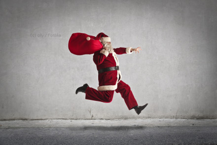 Ein Mann verkleidet als Weihnachtsmann springt in der Luft und trägt einen großen roten Sack auf der Schulter. Der Hintergrund ist eine einfache graue Wand.