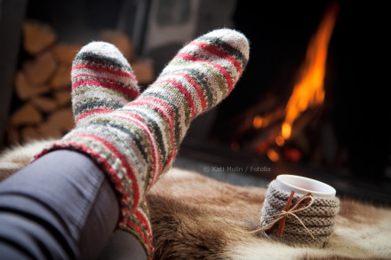 Ein Paar Füße in bunt gestreiften Wollsocken vor einem Kaminfeuer. Im Vordergrund liegt eine Tasse Tee oder Kaffee mit einem gestrickten Überzug auf einem Fell. Im Hintergrund sind Holzscheite zu sehen.