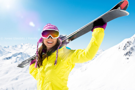 Frau in bunter Winterkleidung trägt Ski auf ihrer Schulter und lächelt in die Kamera. Sie trägt eine pink gestreifte Mütze, eine gelbe Skijacke, Skibrille und Handschuhe. Im Hintergrund sind schneebedeckte Berge und ein klarer, blauer Himmel zu sehen.