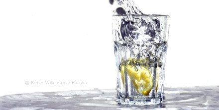 Ein Glas Wasser mit einer Zitronenscheibe, in das Wasser gegossen wird. Das Wasser spritzt und erzeugt Bewegung im Glas.
