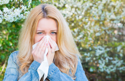 Eine blonde Frau, die im Freien steht und ein Taschentuch an ihre Nase hält, als ob sie niest oder sich die Nase putzt. Im Hintergrund sind blühende Bäume zu sehen.