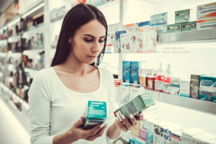 Eine junge Frau steht in einer Apotheke und hält zwei Medikamentenschachteln in den Händen, während sie diese aufmerksam betrachtet. Im Hintergrund sind zahlreiche weitere Medikamentenschachteln auf Regalen zu sehen.