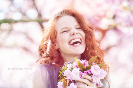 Eine rothaarige Frau lacht herzhaft und hält einen Strauß pinker Blumen in den Händen. Im Hintergrund sind verschwommene, blühende Bäume zu sehen.