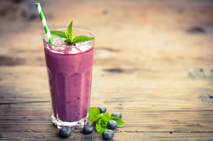 Ein Glas Blaubeer-Smoothie mit einem grünen Papierstrohhalm und frischer Minze als Garnitur. Neben dem Glas befinden sich frische Blaubeeren und Minzeblätter auf einer hölzernen Oberfläche.