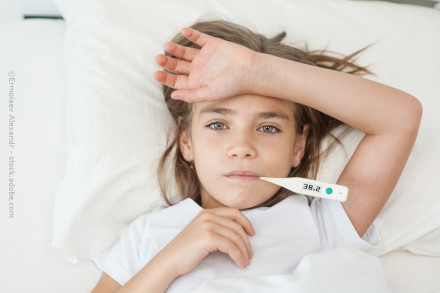 Ein junges Mädchen liegt im Bett. Sie hat ein Thermometer im Mund, das eine Temperatur von 38,2 Grad Celsius anzeigt. Ihre Hand liegt auf ihrer Stirn, während sie erschöpft aussieht. Sie trägt ein weißes T-Shirt und liegt auf einem weißen Kissen.