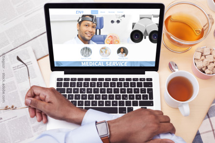 Ein Laptop zeigt eine Medizin-Website mit einem lächelnden Arzt auf dem Bildschirm. Eine Person hält eine Brille in der linken Hand und gibt mit der rechten Hand die Tastatur ein.