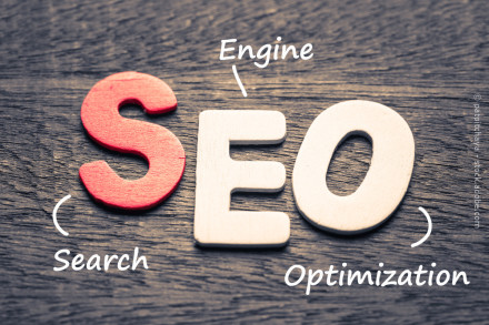 Holzbuchstaben auf einem dunklen Holztisch, die das Wort "SEO" bilden. Das "S" ist rot, während das "E" und das "O" weiß sind. Um die Buchstaben stehen die Wörter "Search", "Engine" und "Optimization" in weißer Schrift, die das Akronym SEO erklären.