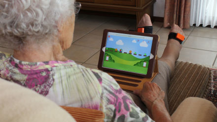 Eine ältere Person mit grauem Haar sitzt auf einem Sofa und hält ein Tablet. Auf dem Tablet-Bildschirm ist ein buntes Videospiel mit einer grünen Wiesenlandschaft und Wolken im Hintergrund zu sehen.
