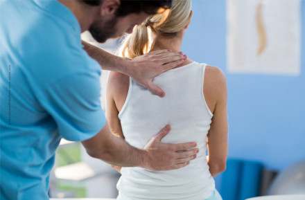 Ein Physiotherapeut in einem blauen Hemd massiert den Rücken einer Frau mit blonden Haaren, die ein weißes ärmelloses Top trägt. Die Frau sitzt und der Physiotherapeut steht hinter ihr, während er ihre Schultern und den unteren Rückenbereich bearbeitet.