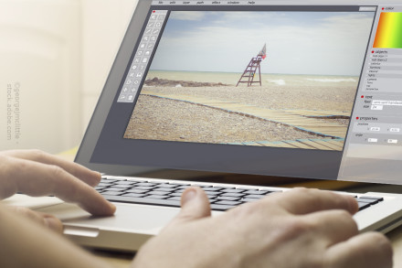 Eine Person arbeitet an einem Laptop, auf dem ein Bildbearbeitungsprogramm geöffnet ist. Auf dem Bildschirm ist ein bearbeitetes Foto von einem leeren Strand mit einem Rettungsschwimmerturm zu sehen. Der Strand hat Kieselsteine und Holzstegwege.