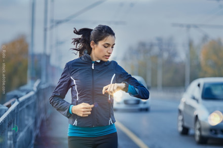 Eine Frau joggt auf einer Straße und schaut auf ihre Armbanduhr. Sie trägt eine schwarze Jacke und ein blaues Sportoberteil. Im Hintergrund sind Autos und leicht verschwommene Bäume zu sehen.