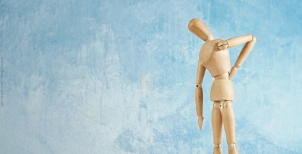 Eine Holzfigur, die Rückenschmerzen darzustellen scheint, steht vor einem blauen Hintergrund. Die Figur hat eine Hand auf den Rücken gelegt und beugt sich leicht nach vorne.