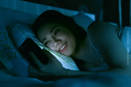 Eine Frau liegt im Bett und lächelt, während sie auf ihr Smartphone schaut. Das Licht vom Bildschirm beleuchtet ihr Gesicht in der dunklen Umgebung.