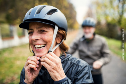 Eine ältere Frau mit einem grauen Fahrradhelm lacht und befestigt den Riemen ihres Helms. Im Hintergrund ist ein älterer Mann, ebenfalls mit Helm, unscharf zu sehen. Die beiden befinden sich auf einem Weg im Freien, umgeben von grüner Natur.