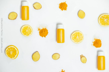Auf einem weißen Hintergrund sind drei kleine, gelbe Flaschen mit weißem Deckel gleichmäßig verteilt. Dazwischen liegen Scheiben von Zitronen, Ingwerstücke und Häufchen von gelbem Kurkumapulver.