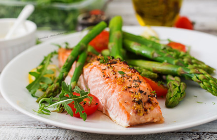 Ein Teller mit gebratenem Lachsfilet, garniert mit Kräutern und Senfkörnern, serviert mit grünem Spargel, Tomaten und Rucola. Im Hintergrund sind eine Schüssel mit Salat und eine Flasche Öl zu sehen.