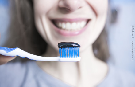 Nahaufnahme eines lächelnden Gesichts, das eine Zahnbürste mit blauen Borsten und schwarzer Zahnpasta darauf hält.