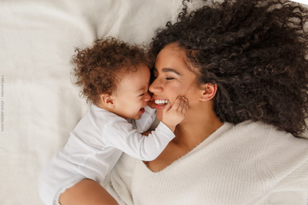 Eine Frau liegt auf einem weißen Bett und hält ein Baby, das ebenfalls weiße Kleidung trägt. Beide lächeln sich liebevoll an und haben lockiges, dunkles Haar. Das Baby berührt das Gesicht der Frau.