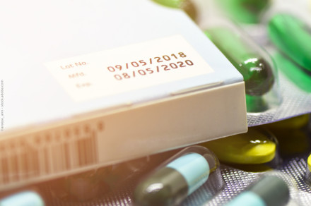 Nahaufnahme einer Medikamentenschachtel mit dem Herstellungsdatum "09/05/2018" und dem Verfallsdatum "08/05/2020". Umliegend sind Blisterpackungen mit grünen, gelben und blau-grünen Kapseln und Tabletten zu sehen.