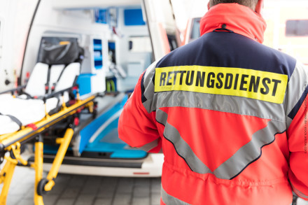 Ein Rettungssanitäter in einer rot-grauen reflektierenden Jacke mit der Aufschrift "RETTUNGSDIENST" auf dem Rücken steht vor einem geöffneten Krankenwagen. Im Hintergrund ist eine Trage sichtbar. Die Szene findet bei Tageslicht statt.