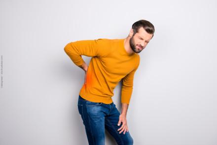 Ein Mann im gelben Pullover und blauer Jeans hat Schmerzen im unteren Rückenbereich. Er hält seine Hand auf die schmerzende Stelle und verzieht das Gesicht vor Schmerzen vor einem weißen Hintergrund.