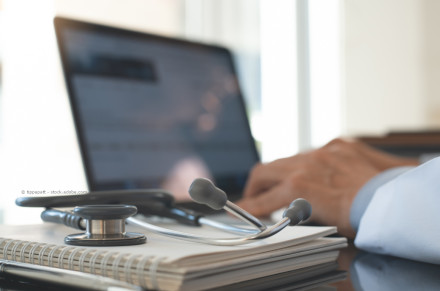 Ein Stethoskop liegt auf einem Notizbuch, während im Hintergrund die Hände einer Person auf einer Computertastatur tippen.