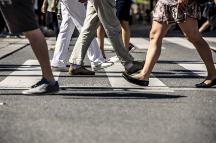 Menschen überqueren einen Zebrastreifen in einer Stadt. Nur die Beine und Füße der Personen sind sichtbar; sie tragen verschiedene Arten von Schuhen und Kleidung. Der Asphalt und die weißen Streifen des Zebrastreifens sind deutlich zu erkennen.