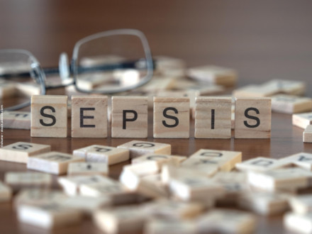 Auf dem Bild sind Holzbuchstabenblöcke zu sehen, die das Wort "SEPSIS" bilden. Die Blöcke liegen auf einem Holztisch. Im Hintergrund sind andere Buchstabensteine verstreut und eine Brille ist leicht unscharf zu erkennen.