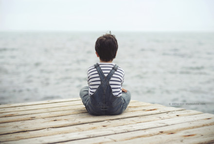 Ein Kind in gestreiftem Langarmshirt und blauer Latzhose sitzt auf einem Holzsteg. Das Kind ist dem Betrachter den Rücken zugewandt und schaut auf das Meer hinaus.