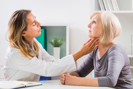 Eine Ärztin untersucht den Hals einer älteren Patientin in einem hellen, modernen Behandlungsraum. Die Ärztin trägt einen weißen Kittel und die Patientin ein graues Oberteil.