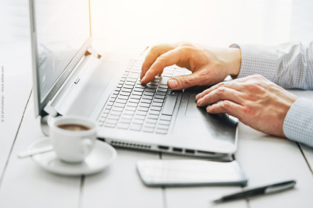 Nahaufnahme von Händen, die auf einem Laptop-Tastatur tippen. Auf dem Tisch stehen eine weiße Kaffeetasse mit Untertasse und Löffel, ein Stift und ein Smartphone. Sanftes Sonnenlicht fällt von oben links auf die Szene. Eine helle, produktive Atmosphäre.