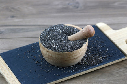 Ein Holzmörser und -stößel gefüllt mit Chiasamen steht auf einem schwarzen Schieferbrett, das auf einem Holzschneidebrett liegt. Einige Chiasamen sind um den Mörser herum verstreut. Der Hintergrund besteht aus einer holzartigen Oberfläche.