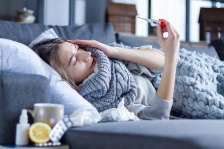 Eine Frau liegt krank auf einem Sofa, eingehüllt in eine graue Decke und einen Schal. Sie hält sich den Kopf und schaut auf ein Thermometer in ihrer Hand.