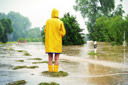 Eine Person steht im Regen auf einer überschwemmten Straße, gekleidet in einem gelben Regenmantel und gelben Gummistiefeln. Umgeben von hochwasserführendem Wasser und grüner Vegetation im Hintergrund.