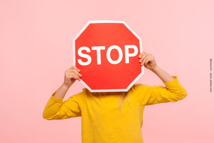 Eine Person in einem gelben Pullover hält ein großes rotes STOP-Schild vor ihrem Gesicht. Der Hintergrund ist rosa.