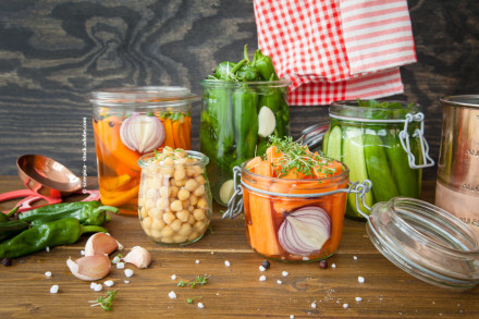Glasgefäße mit eingelegtem Gemüse auf einem Holztisch; enthaltene Zutaten sind marinierte Karotten, Kichererbsen, grüne Paprika und Gurken. Einige Knoblauchzehen, grobkörniges Salz und eine rot-weiß karierte Stoffserviette sind ebenfalls zu sehen.